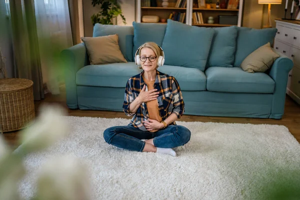 一位成熟的高年级女性使用耳机进行在线引导冥想练习心神瑜珈 在家闭目养神真正的人自我护理概念复制空间 — 图库照片