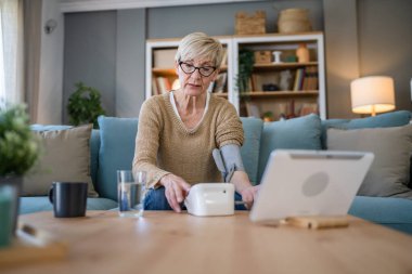 Üst düzey beyaz bir kadın, evde oturup gerçek sağlık hizmetleri kavramını incelerken, sağlık sonuçlarını kontrol etmek için elinde tansiyon aleti kullanıyor.