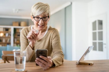Kafkasyalı yaşlı bir kadın ilaç ya da vitamin takviyesi için evde bir bardak su içen bir kadın internet üzerinden gerçek insanların sağlık hizmetleri konseptini arıyor.