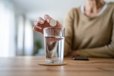 Yaşlı bir beyaz kadın ilaç ya da vitamin takviyesi hapı alıyor. Bir bardak suya koyuyor. Gerçek sağlık hizmetleri konseptinde tedaviyi hazırlıyor.