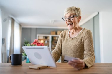 Yetişkin bir yaşlı kadın büyükanne evde oturup online alışveriş siteleri için kredi kartı ya da banka kartı kullanıyor. Bir şeyler satın alıyor. Dijital tablet kullanıyor. Gerçek insanlar yer kopyalıyor.