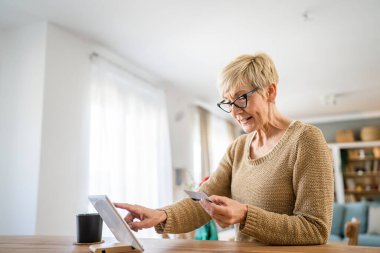 Yetişkin bir yaşlı kadın büyükanne evde oturup online alışveriş siteleri için kredi kartı ya da banka kartı kullanıyor. Bir şeyler satın alıyor. Dijital tablet kullanıyor. Gerçek insanlar yer kopyalıyor.