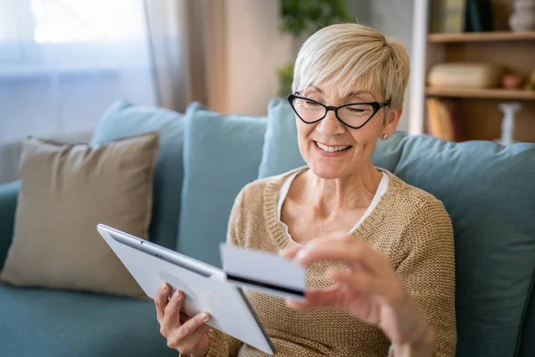 ある成熟した年配の女性の祖母はオンラインショッピングのための家の使用のクレジットカードかデビットカードに坐るインターネットの店物を買うことはデジタルタブレットを使用する実際の人々はスペースをコピーする — ストック写真