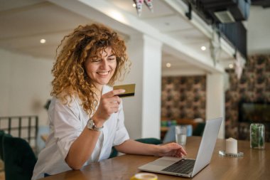 Kıvırcık saçlı, mutlu, yetişkin bir kadın Kafe 'deki laptop bilgisayarından kredi kartı alışverişi yapıyor. Gerçek insanlar e-bankacılık internet indirimi yapıyor.