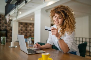 Kıvırcık saçlı, mutlu, yetişkin bir kadın Kafe 'deki laptop bilgisayarından kredi kartı alışverişi yapıyor. Gerçek insanlar e-bankacılık internet indirimi yapıyor.