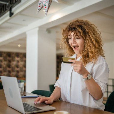 Kıvırcık saçlı, mutlu, yetişkin bir kadın Kafe 'deki laptop bilgisayarından kredi kartı alışverişi yapıyor. Gerçek insanlar e-bankacılık internet indirimi yapıyor.