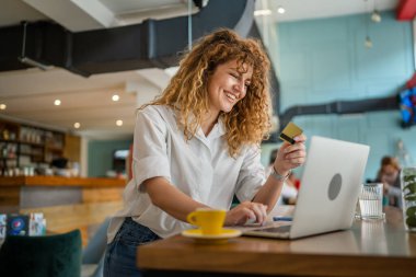 Kıvırcık saçlı, mutlu, yetişkin bir kadın Kafe 'deki laptop bilgisayarından kredi kartı alışverişi yapıyor. Gerçek insanlar e-bankacılık internet indirimi yapıyor.