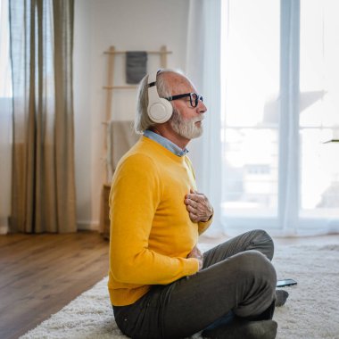 Yetişkin bir beyaz erkek internet rehberli meditasyon için kulaklık kullanıyor evde gözleri kapalı yoga tezahürü yapıyor gerçek insanların kişisel bakım kavramını taklit ediyor.