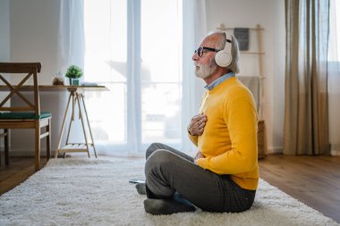 Yetişkin bir beyaz erkek internet rehberli meditasyon için kulaklık kullanıyor evde gözleri kapalı yoga tezahürü yapıyor gerçek insanların kişisel bakım kavramını taklit ediyor.