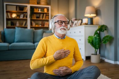 Yetişkin bir beyaz erkek internet rehberli meditasyon için kulaklık kullanıyor evde gözleri kapalı yoga tezahürü yapıyor gerçek insanların kişisel bakım kavramını taklit ediyor.