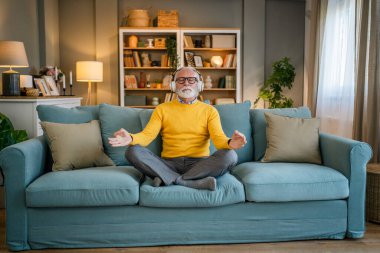 Yetişkin bir beyaz erkek internet rehberli meditasyon için kulaklık kullanıyor evde gözleri kapalı yoga tezahürü yapıyor gerçek insanların kişisel bakım kavramını taklit ediyor.