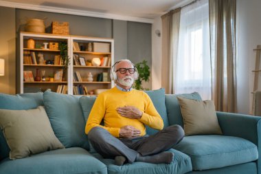 Yetişkin bir beyaz erkek internet rehberli meditasyon için kulaklık kullanıyor evde gözleri kapalı yoga tezahürü yapıyor gerçek insanların kişisel bakım kavramını taklit ediyor.