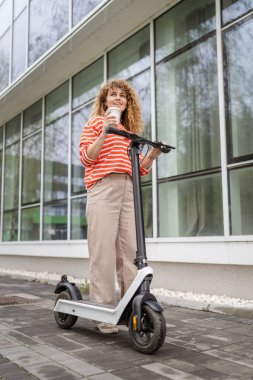 Bir kadın yetişkin beyaz kadın elektrik tekmeli scooter ile şehirde ayakta duruyor. Gündüz güvenli modern alternatif ulaşım aracı fotokopi alanı.