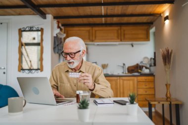 Son sınıf beyaz erkek büyükbaba evinde oturup ağrı kesici veya vitamin takviyesi paketini tutuyor. Bilgisayardan sipariş edilen dizüstü bilgisayarın yanında fotokopi odası almaya hazır.
