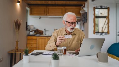 Son sınıf beyaz erkek büyükbaba evinde oturup ağrı kesici veya vitamin takviyesi paketini tutuyor. Bilgisayardan sipariş edilen dizüstü bilgisayarın yanında fotokopi odası almaya hazır.