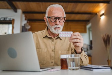 Son sınıf beyaz erkek büyükbaba evinde oturup ağrı kesici veya vitamin takviyesi paketini tutuyor. Bilgisayardan sipariş edilen dizüstü bilgisayarın yanında fotokopi odası almaya hazır.