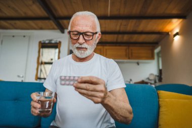 Son sınıf beyaz erkek büyükbaba evinde oturup ağrı kesici ya da vitamin takviyesi paketini tutuyor. İlaç almaya hazır. Gerçek insan kopyalama alanı.