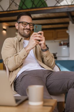 Yetişkin bir beyaz adam evinde oturup video oyunu oynar ya da mesajlaşır, boş zaman aktivitesi yapar cep telefonu fotokopi alanı, gözlük ve gömlek giyer.