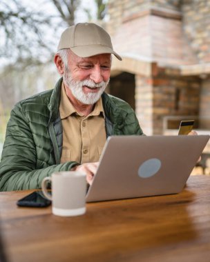 Sakallı, kıdemli, beyaz bir erkek bütün gün dışarıda dizüstü bilgisayarla alışveriş yapıyor. Bir şeyler alıyor, kredi kartı kullanıyor. Emekliliğinin tadını çıkarıyor.