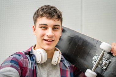 Genç, yetişkin, beyaz, kaykaycı bir genç omuzunda kaykayla ve kulaklıklarıyla kameraya bakıp mutlu bir şekilde kendine güvenen gömleğini giyer ve gerçek uzay fotokopisi çeker.