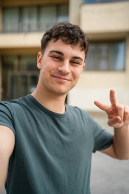 Genç, yetişkin, beyaz, açık havada kendi portresi pozu veren bir genç, kameraya bakıyor. Mutlu, kendine güvenen, sıradan bir gömlek giyiyor. Uzaydaki çirkin kullanıcının ürettiği içeriği kopyalıyor.