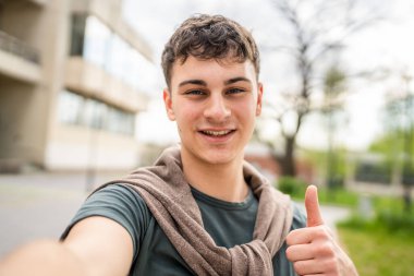 Genç, yetişkin, beyaz, açık havada kendi portresi pozu veren bir genç, kameraya bakıyor. Mutlu, kendine güvenen, sıradan bir gömlek giyiyor. Uzaydaki çirkin kullanıcının ürettiği içeriği kopyalıyor.