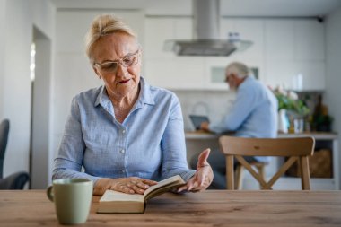 Yetişkin, yaşlı, Kafkasyalı, emekli kadın, modern büyükanne evde kitap okuyor, gözlük takıyor, kocası ise aile yaşamının arkasında duruyor.