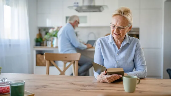 一位成熟的老年女性退休老人现代祖母戴着眼镜在家里看书 而她的丈夫则站在家庭生活的后面积极的老年人模仿空间 — 图库照片