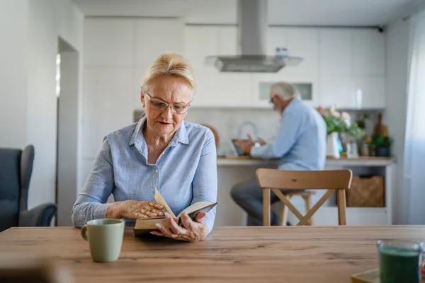 一位成熟的老年女性退休老人现代祖母戴着眼镜在家里看书 而她的丈夫则站在家庭生活的后面积极的老年人模仿空间 — 图库照片