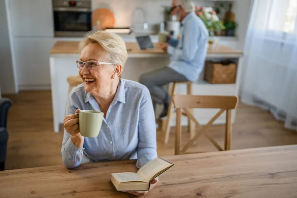 一人の女性成熟したシニア白人女性年金現代の祖母読みます本で家庭用眼鏡で彼女の夫は家族生活の後ろに立っている間アクティブシニアリアル人々コピースペース — ストック写真