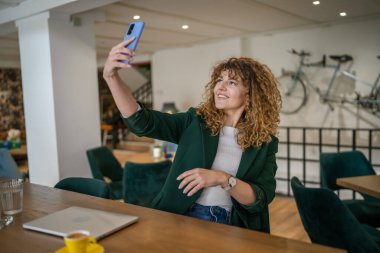 Kafe 'de oturan yetişkin bir kadın internet görüntülü konuşma ya da kendi portresi için akıllı telefon kullanıyor gülümseyen gerçek bir insana iyi haberler veren mutlu bir gülümseme.