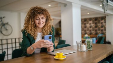 Kafe 'de oturan yetişkin bir kadın internet gezintisi ya da mesaj atmak için akıllı telefon kullanıyor. Mutlu bir gülümseme, güler yüzlü gerçek bir insan gibi gülümsüyor.