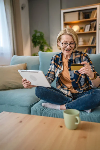 Yetişkin bir yaşlı kadın büyükanne evde oturup online alışveriş siteleri için kredi kartı ya da banka kartı kullanıyor. Bir şeyler satın alıyor. Dijital tablet kullanıyor. Gerçek insanlar yer kopyalıyor.