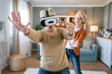 İki olgun kadın Kafkasyalı kadın arkadaş veya kız kardeş evde sanal gerçekliğin tadını çıkarıyorlar. VR kulaklığı. Son sınıf öğrencileri eğleniyor.
