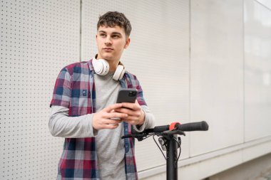 Bir genç beyaz erkek, elektrik tekmeli genç Z nesli şehir eko taşıma konseptindeki modern binada scooter 'ı itiyor SMS mesajları için cep telefonu kullanıyor.