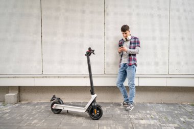 Bir genç beyaz erkek, elektrik tekmeli genç Z nesli şehir eko taşıma konseptindeki modern binada scooter 'ı itiyor SMS mesajları için cep telefonu kullanıyor.