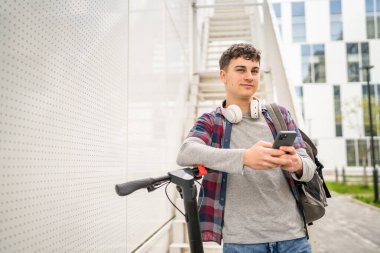Bir genç beyaz erkek, elektrik tekmeli genç Z nesli şehir eko taşıma konseptindeki modern binada scooter 'ı itiyor SMS mesajları için cep telefonu kullanıyor.
