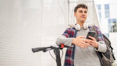 Bir genç beyaz erkek, elektrik tekmeli genç Z nesli şehir eko taşıma konseptindeki modern binada scooter 'ı itiyor SMS mesajları için cep telefonu kullanıyor.