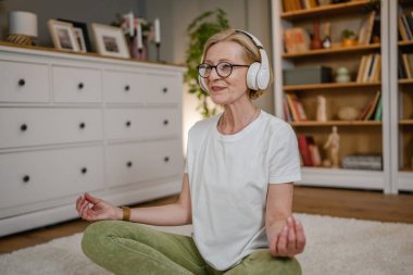 Yetişkin bir yaşlı beyaz kadın internet rehberli meditasyon için kulaklık kullanıyor. Farkındalık yogası yapıyor. Evde gözleri kapalı.