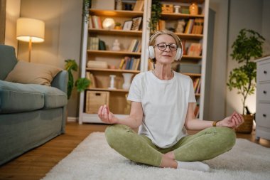 Yetişkin bir yaşlı beyaz kadın internet rehberli meditasyon için kulaklık kullanıyor. Farkındalık yogası yapıyor. Evde gözleri kapalı.