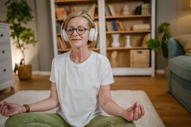Yetişkin bir yaşlı beyaz kadın internet rehberli meditasyon için kulaklık kullanıyor. Farkındalık yogası yapıyor. Evde gözleri kapalı.