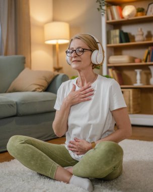 Yetişkin bir yaşlı beyaz kadın internet rehberli meditasyon için kulaklık kullanıyor. Farkındalık yogası yapıyor. Evde gözleri kapalı.