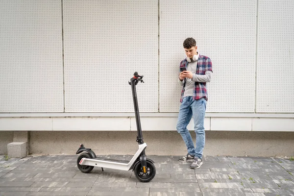 交通概念の都市エコモードで近代的な建物で電動キックプッシュスクーターと一人の若い白人男性十代の世代Zは ミリ秒のテキストメッセージコピースペースのための携帯電話を使用しています — ストック写真