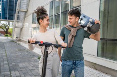 Genç adam ve kadın genç çift, beyaz erkek ve kız kardeş, mutlu bir gülümsemeyle modern binanın önünden geçiyorlar. Kaykay ve elektrikli scooter taşıyorlar.