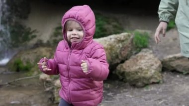 Küçük beyaz bir kız çocuğu, iki yaşında, kış veya sonbahar günlerinde doğada kameraya doğru koşuyor, ceketini giyiyor, çocukluğunu yavaş çekimde büyütüyor, mutluluk fotokopisi çekiyor.