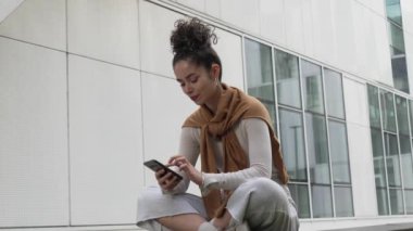 Bir yetişkin kadın beyaz kız öğrenci üniversitenin ya da modern binanın önünde dışarıda oturuyor cep telefonu akıllı telefonu kullanıyor mesajlar gönderiyor ya da interneti yavaş çekimde tarıyor
