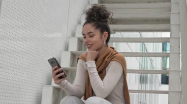 Bir yetişkin kadın beyaz kız öğrenci üniversitenin ya da modern binanın önünde dışarıda oturuyor cep telefonu akıllı telefonu kullanıyor mesajlar gönderiyor ya da interneti yavaş çekimde tarıyor