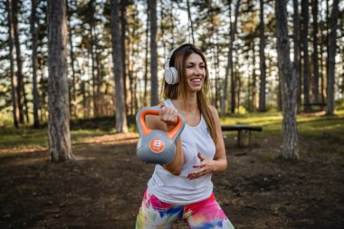 One woman adult caucasian female athlete using kettlebell girya weight during training in the forest park woods in summer day with headphones happy smile brunette health and fitness concept copy space