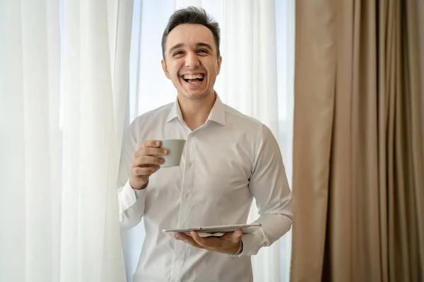 stock image One man adult caucasian male standing by the window at home office or hotel room with cup of coffee and digital tablet taking a brake from work and preparing real people copy space wear white shirt