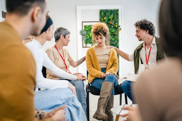 A group of people sitting in a circle with two therapists and a woman in the center discussing and providing emotional support for her The focus is on the woman but everyone is engaged in conversation
